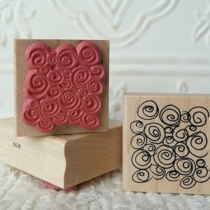 May include: Two wooden rubber stamps with different designs. The first stamp has a red swirl pattern and the second stamp has a black swirl pattern. The stamps are both square and have a wooden base.