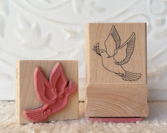 Op de afbeelding: Twee houten stempels met een duivenontwerp. Een stempel heeft een rode duif met een olijftak in zijn bek, terwijl de andere stempel een zwarte omtrek van een duif met een olijftak in zijn bek heeft.
