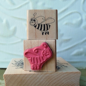 May include: A set of three wooden stamps featuring a bee and a butterfly design. The stamps are stacked on top of each other and are ready to be used for crafting projects.