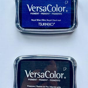 May include: Two ink pads, one blue and one black, with the brand name "VersaColor" and the text "PIGMENT PIGMENT PIGMENTO" printed on the front. The blue ink pad says "Royal Blue / Bleu Royal / Azul real" and the black ink pad says "Pinecone / Pomme de Pin / Marr&oacute;n pi&ntilde;a". Both ink pads have the brand name "TSUKINEKO&reg;" printed on the bottom.