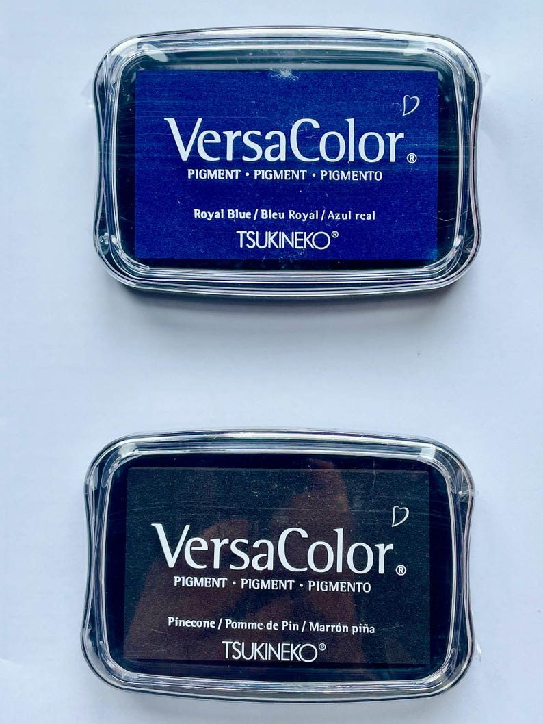 May include: Two ink pads, one blue and one black, with the brand name "VersaColor" and the text "PIGMENT PIGMENT PIGMENTO" printed on the front. The blue ink pad says "Royal Blue / Bleu Royal / Azul real" and the black ink pad says "Pinecone / Pomme de Pin / Marr&oacute;n pi&ntilde;a". Both ink pads have the brand name "TSUKINEKO&reg;" printed on the bottom.