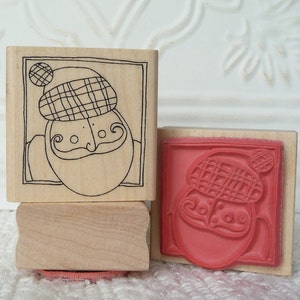 May include: Two wooden rubber stamps with a cartoon image of a person wearing a plaid hat. The top stamp is in black and white and the bottom stamp is in red.