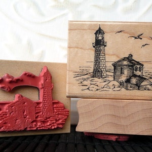 Puede incluir: Tres sellos de goma montados en madera con diseños de faros. El sello superior presenta una ilustración en blanco y negro de un faro en una costa rocosa con pájaros volando por encima. El sello del medio presenta una ilustración roja y blanca de un faro con un edificio al lado. El sello inferior está en blanco.