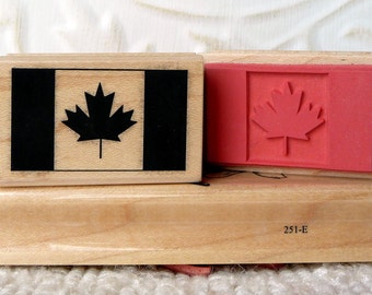 Pode incluir: Dois carimbos de madeira com desenhos de bandeiras canadenses. O carimbo superior apresenta uma bandeira canadense em preto e branco com uma folha de bordo. O carimbo inferior apresenta uma bandeira canadense vermelha com uma folha de bordo. Os carimbos estão empilhados em um bloco de madeira com o número 251-E impresso nele.