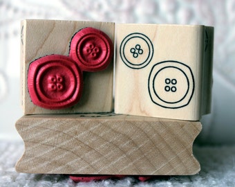 May include: Two wooden blocks with red and black button designs. The top block has two red buttons with four holes each. The bottom block has a black button with four holes and a black button with six holes.