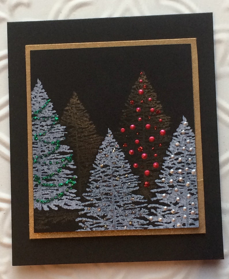 May include: A Christmas card with a black background and a gold border. The card depicts a forest of stylized evergreen trees in shades of grey, brown, and white, some decorated with red and green embellishments. The design creates a festive winter scene.
