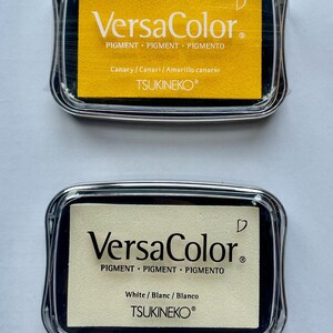 May include: Two ink pads, one yellow and one white, with the text "VersaColor" and "TSUKINEKO" on them. The ink pads are in clear plastic cases.