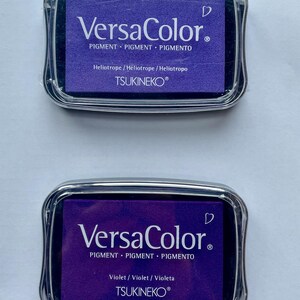 May include: Two ink pads for stamping, both are purple. The top pad is labeled "VersaColor" and "Heliotrope/H&eacute;liotrope / Heliotropo". The bottom pad is labeled "VersaColor" and "Violet/Violet / Violeta". Both pads are labeled "TSUKINEKO&reg;".