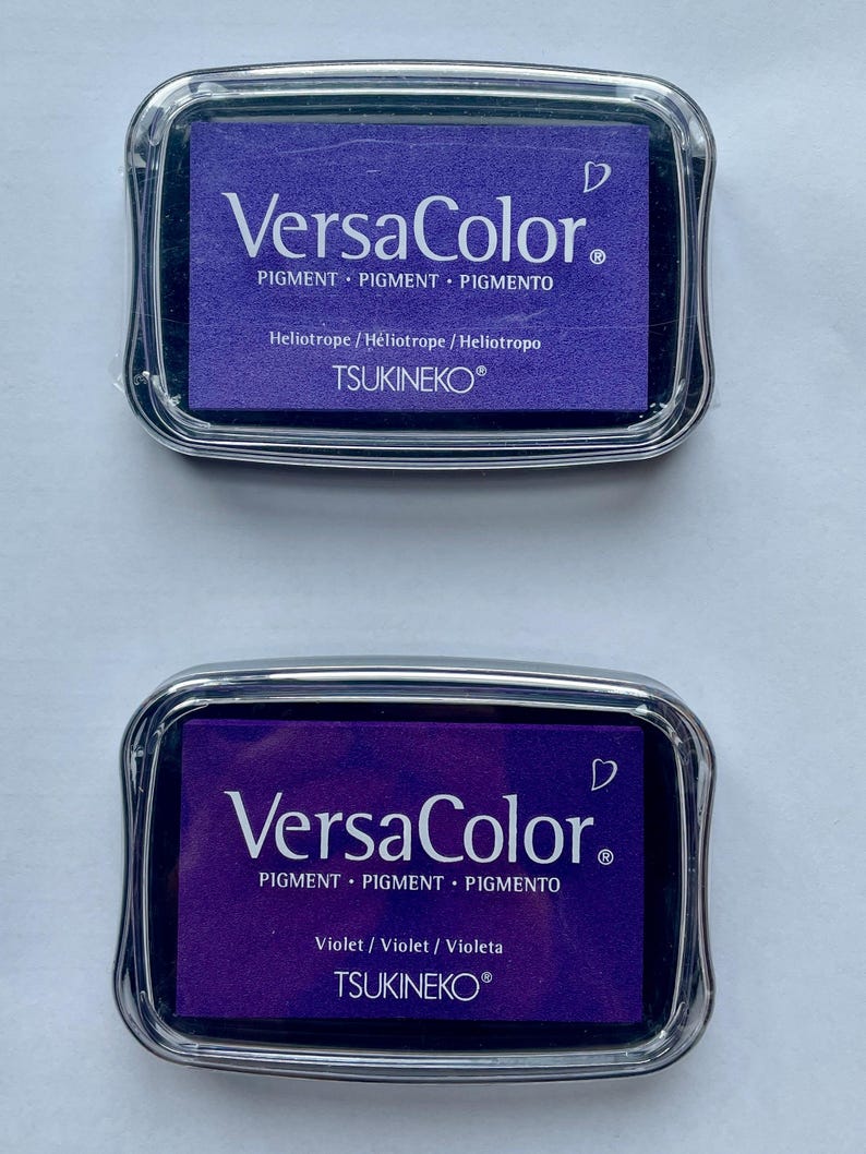 May include: Two ink pads for stamping, both are purple. The top pad is labeled "VersaColor" and "Heliotrope/H&eacute;liotrope / Heliotropo". The bottom pad is labeled "VersaColor" and "Violet/Violet / Violeta". Both pads are labeled "TSUKINEKO&reg;".