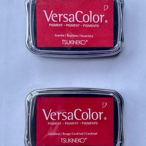 May include: Two red ink pads with black lids. The ink pads are labeled "VersaColor" and have the text "PIGMENT PIGMENT PIGMENTO" and "TSUKINEKO&reg;" printed on them. The top ink pad is labeled "Scarlet/Ecarlate/Escarlata" and the bottom ink pad is labeled "Cardinal/Rouge Cardinal/Cardinal".