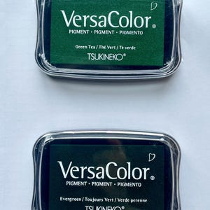 May include: Two ink pads for stamping, both are green. The top pad is labeled "VersaColor" with the text "Green Tea / The Vert / T&eacute; verde" and "TSUKINEKO&reg;". The bottom pad is labeled "VersaColor" with the text "Evergreen/Toujours Vert / Verde perenne" and "TSUKINEKO&reg;".