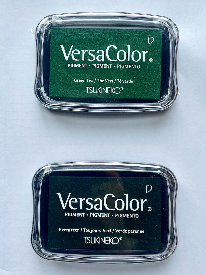 May include: Two ink pads for stamping, both are green. The top pad is labeled "VersaColor" with the text "Green Tea / The Vert / T&eacute; verde" and "TSUKINEKO&reg;". The bottom pad is labeled "VersaColor" with the text "Evergreen/Toujours Vert / Verde perenne" and "TSUKINEKO&reg;".