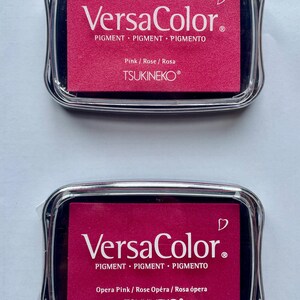 May include: Two pink ink pads with black lids. The top pad says "VersaColor" and "Pink/Rose/Rosa" and the bottom pad says "VersaColor" and "Opera Pink/Rose Op&eacute;ra/Rosa &oacute;pera". Both pads have the text "PIGMENT PIGMENT PIGMENTO" and "TSUKINEKO&reg;".