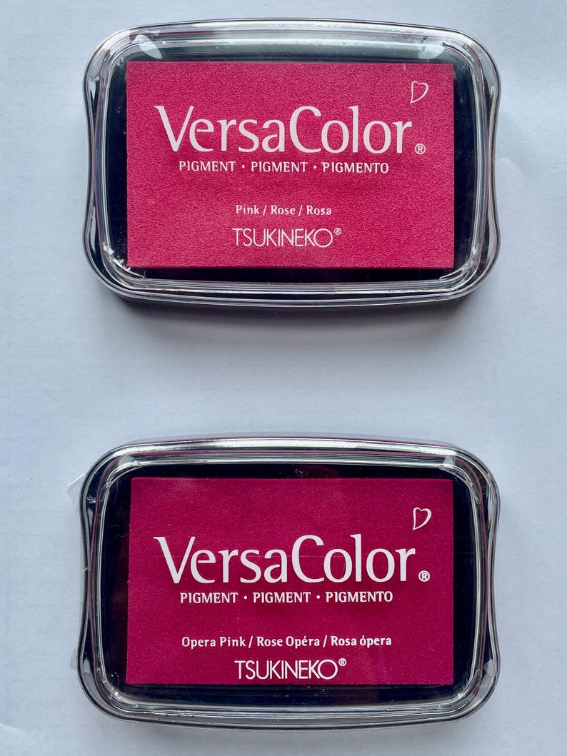 May include: Two pink ink pads with black lids. The top pad says "VersaColor" and "Pink/Rose/Rosa" and the bottom pad says "VersaColor" and "Opera Pink/Rose Op&eacute;ra/Rosa &oacute;pera". Both pads have the text "PIGMENT PIGMENT PIGMENTO" and "TSUKINEKO&reg;".