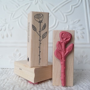 Puede incluir: Dos sellos de madera con diseños de rosas. Un sello presenta un dibujo en línea negra de una rosa con tallo y hojas. El otro sello presenta un diseño en relieve rojo de una rosa con tallo y hojas. El texto en el sello dice "Salt Spring Island, BC, Canada".