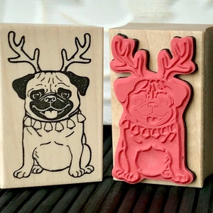 May include: Two wooden rubber stamps featuring a pug wearing reindeer antlers. The stamp on the left is black and white, while the stamp on the right is red.