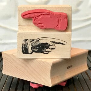以下が含まれることがあります： 指を指している木製のゴム印が2つ。上の印はピンク色の指が右を指している。下の印は白黒の指が右を指している。印の下には1047-Cという番号が書かれている。