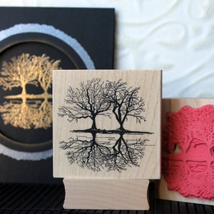 May include: Three wooden blocks with images of trees. The top block has a black and white image of two trees reflected in water. The middle block has a black and white image of two trees reflected in water. The bottom block has a red image of two trees reflected in water.