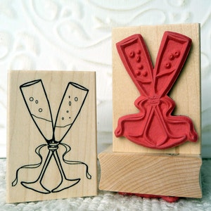 May include: Two wooden blocks with rubber stamps. One stamp features a red silhouette of two champagne flutes tied together with a ribbon. The other stamp features a black line drawing of two champagne flutes with bubbles and a ribbon.