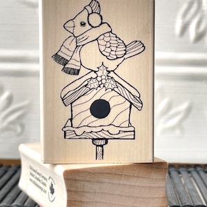 May include: A wooden rubber stamp featuring a black and white illustration of a bird wearing a scarf and earmuffs perched on a birdhouse with holly.