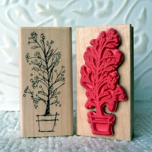 May include: Two wooden stamps featuring a plant design. The stamp on the left has a black outline of a plant with berries in a pot. The stamp on the right has a red raised design of a plant with berries in a pot.