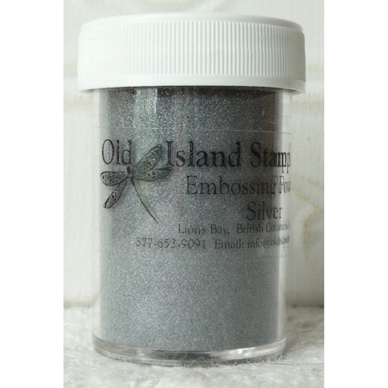 May include: A clear plastic jar filled with silver embossing powder. The jar has a white lid and a black label that reads "Old Island Stamp Embossing Powder Silver".