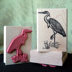 Op de afbeelding: Twee houten blokken met reliëfbeelden van reigers. Het bovenste blok toont een zwart-wit beeld van een reiger die in het water staat. Het onderste blok toont een rood beeld van een reiger die in riet staat.