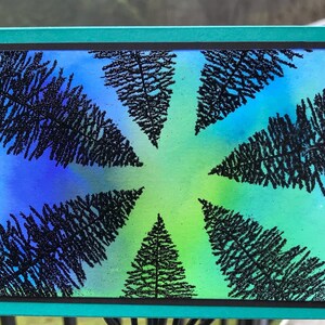 May include: A rectangular artwork with a blue and green gradient background, featuring black silhouettes of pine trees in a circular arrangement. The artwork is framed with a teal border.