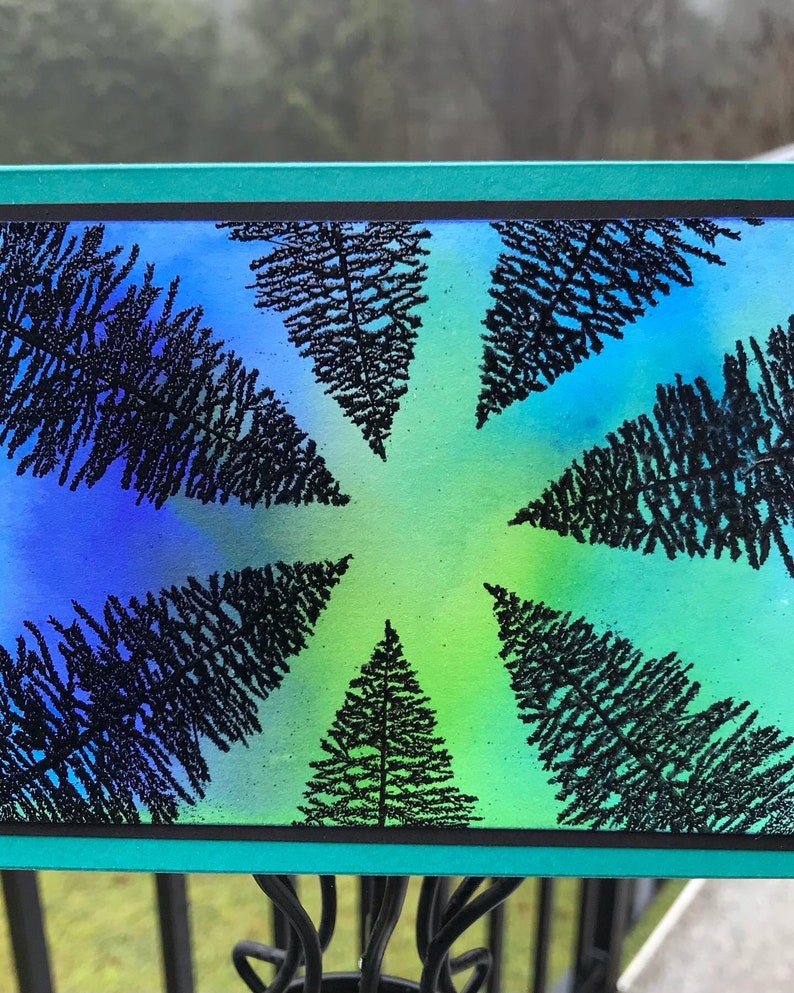 May include: A rectangular artwork with a blue and green gradient background, featuring black silhouettes of pine trees in a circular arrangement. The artwork is framed with a teal border.