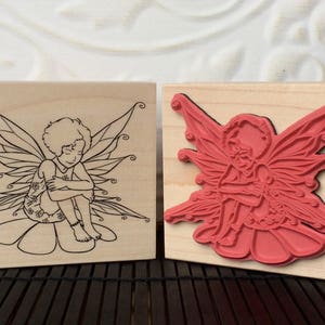 May include: Two square wooden rubber stamps featuring a line art illustration of a person with curly hair and wings sitting on a flower. One stamp shows the black ink design on wood, while the other shows the raised red rubber relief of the same image.