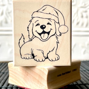 May include: A wooden rubber stamp with a black and white illustration of a puppy wearing a Santa hat. The stamp reads "Artist:  Real  Redhead  1150-A".