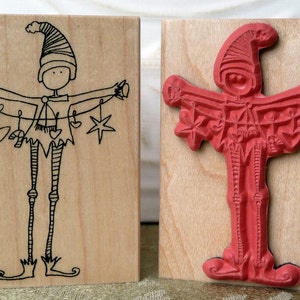 May include: Two wooden stamps featuring a whimsical elf character holding a string of Christmas ornaments. The first stamp is uninked and shows a black line drawing of the elf. The second stamp is inked in red and shows the elf holding a string of ornaments.