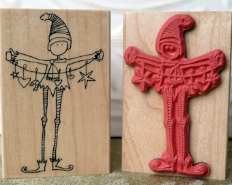 May include: Two wooden stamps featuring a whimsical elf character holding a string of Christmas ornaments. The first stamp is uninked and shows a black line drawing of the elf. The second stamp is inked in red and shows the elf holding a string of ornaments.