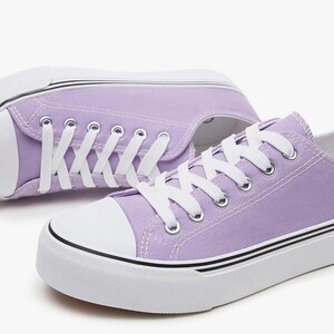 May include: A pair of low-top canvas sneakers in a light lavender color. The shoes feature white rubber toe caps, white laces, and thick white soles with thin black horizontal stripes running along the sides of the rubber midsole base.