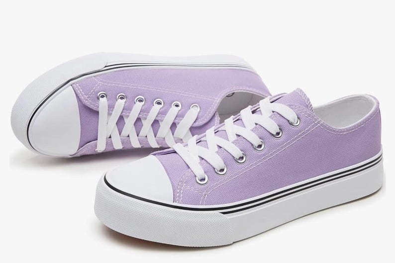 May include: A pair of low-top canvas sneakers in a light lavender color. The shoes feature white rubber toe caps, white laces, and thick white soles with thin black horizontal stripes running along the sides of the rubber midsole base.
