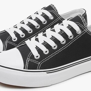 May include: A pair of low-top sneakers featuring a black canvas upper, white laces, and a white rubber toe cap. The shoes have a thick white rubber sole with a thin black stripe detail running along the side, providing a classic athletic look.