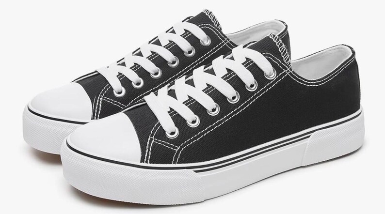May include: A pair of low-top sneakers featuring a black canvas upper, white laces, and a white rubber toe cap. The shoes have a thick white rubber sole with a thin black stripe detail running along the side, providing a classic athletic look.