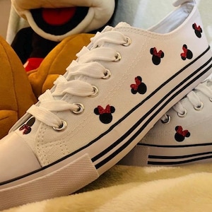 Puede incluir: Zapatillas blancas de lona con ribetes negros y aplicaciones de Minnie Mouse con purpurina roja.