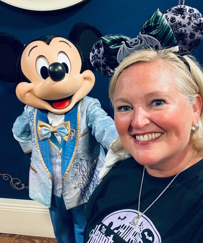 May include: A Mickey Mouse character figure wearing a light blue and gold tuxedo with a matching bow tie, standing next to a person wearing a black headband with two ears featuring a green and black striped pattern, a bat graphic, and a black lace overlay.