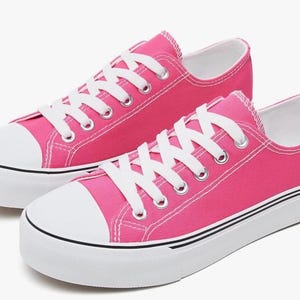 May include: A pair of low-top canvas sneakers in a vibrant hot pink color. The shoes feature white laces, white rubber toe caps, and thick white rubber soles with a thin black stripe detail running along the side of the platform base.