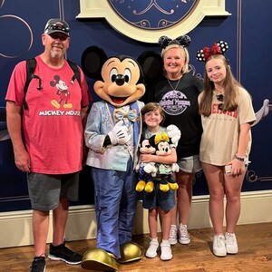 May include: Mickey Mouse in a light blue suit stands with a group of four people. The group includes an adult in a red Mickey Mouse t-shirt, an adult in a black Haunted Mansion t-shirt, a child holding two plush toys, and a teenager in a beige t-shirt.