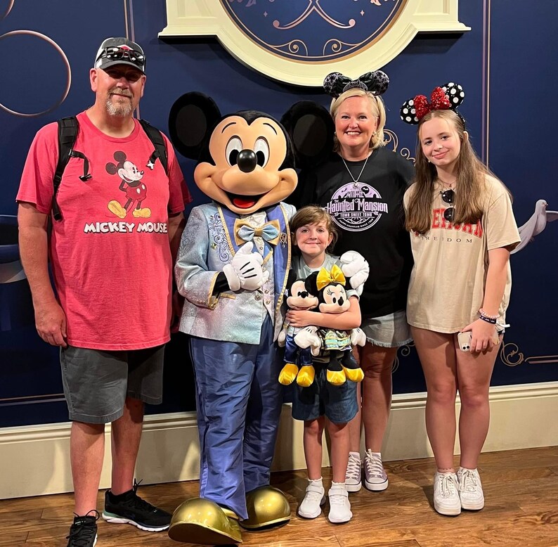 May include: Mickey Mouse in a light blue suit stands with a group of four people. The group includes an adult in a red Mickey Mouse t-shirt, an adult in a black Haunted Mansion t-shirt, a child holding two plush toys, and a teenager in a beige t-shirt.