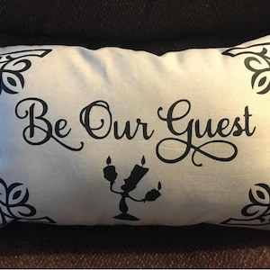Könnte beinhalten: Ein weißes Kissen mit schwarzem Besatz und einer schwarzen Silhouette von Lumière aus Die Schöne und das Biest. Das Kissen trägt die Aufschrift "Be Our Guest" in schwarzer Kursivschrift.