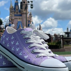 May include: A pair of lavender canvas sneakers featuring a repeating white pattern of small mouse head silhouettes. The low-top shoes have white laces, silver metal eyelets, a white rubber toe cap, and a white rubber sole with a thin black stripe.