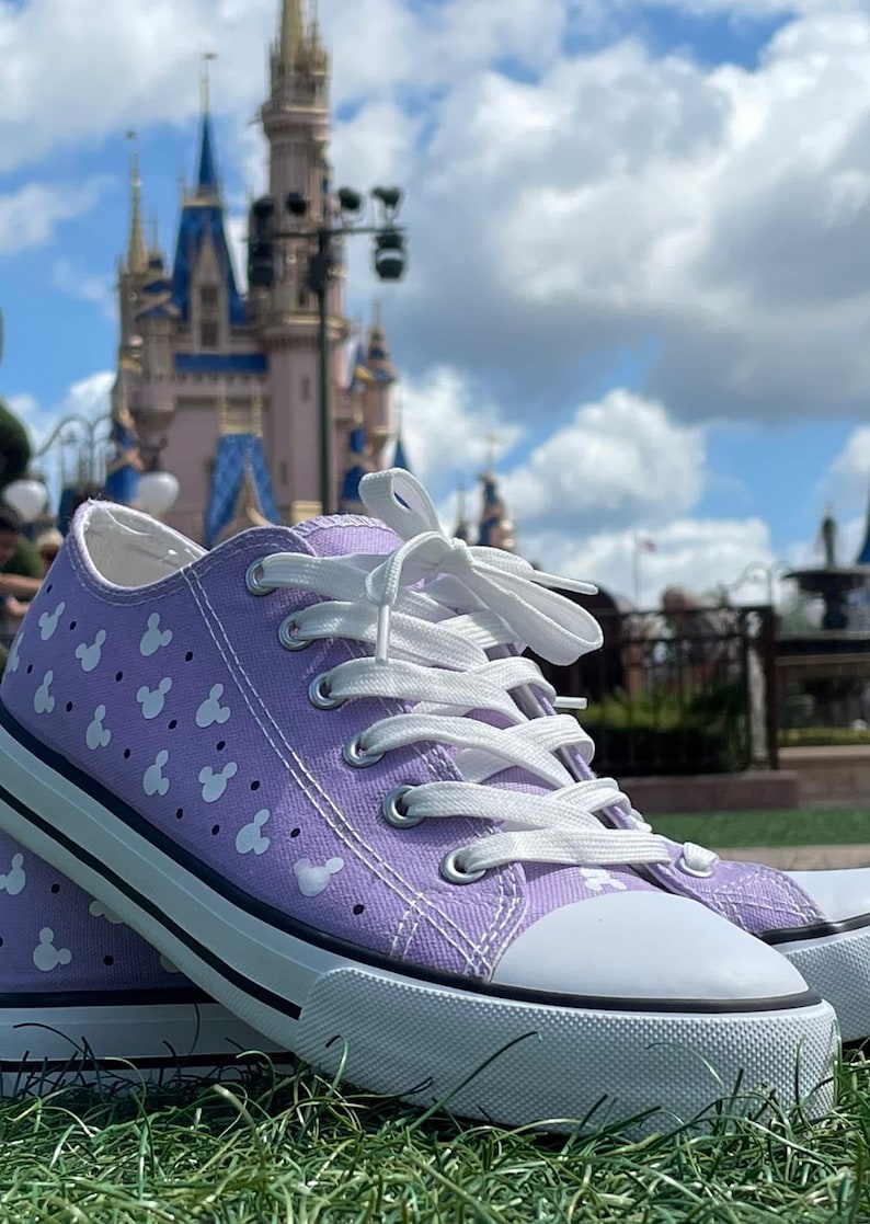 May include: A pair of lavender canvas sneakers featuring a repeating white pattern of small mouse head silhouettes. The low-top shoes have white laces, silver metal eyelets, a white rubber toe cap, and a white rubber sole with a thin black stripe.