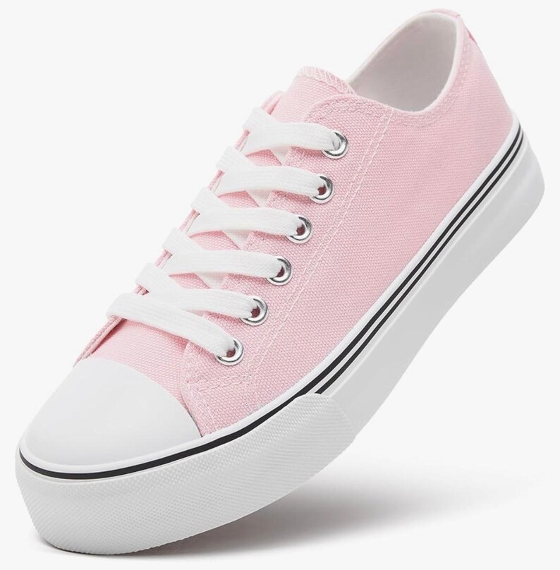 May include: A low-top sneaker featuring a light pink canvas upper, white laces, and silver-toned metal eyelets. The shoe includes a white rubber toe cap and a thick white rubber sole accented with two thin black horizontal stripes along the side.