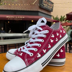 May include: A pair of low-top canvas sneakers in a deep maroon color featuring a repeating white Mickey Mouse head pattern. The shoes have white laces, a white rubber toe cap, and a thick white sole with two thin black horizontal stripes.