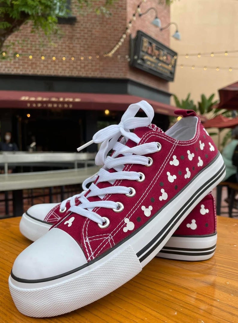 May include: A pair of low-top canvas sneakers in a deep maroon color featuring a repeating white Mickey Mouse head pattern. The shoes have white laces, a white rubber toe cap, and a thick white sole with two thin black horizontal stripes.