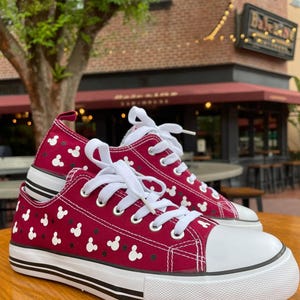 May include: A pair of low-top canvas sneakers in deep red featuring a repeating pattern of small white Mickey Mouse head silhouettes and black dots. The shoes have white laces, white rubber toe caps, and white soles with two thin black horizontal stripes.