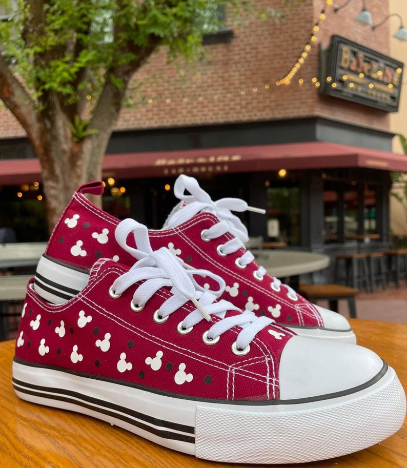 May include: A pair of low-top canvas sneakers in deep red featuring a repeating pattern of small white Mickey Mouse head silhouettes and black dots. The shoes have white laces, white rubber toe caps, and white soles with two thin black horizontal stripes.